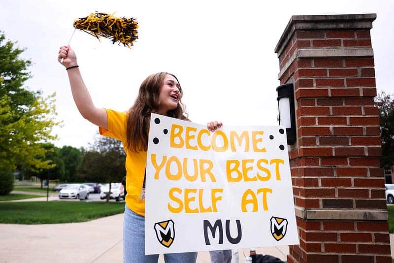 A student waves a pom pom and a banner that reads "Become your best self at MU"